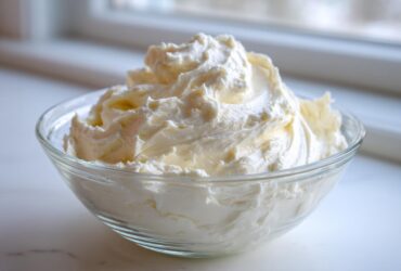 A clear glass bowl filled with perfectly swirled, creamy vanilla buttercream frosting, set near a bright window.