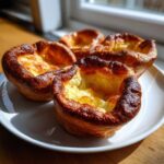 Four golden brown, fluffy yorkshire pudding cups sitting on a white plate near a window.