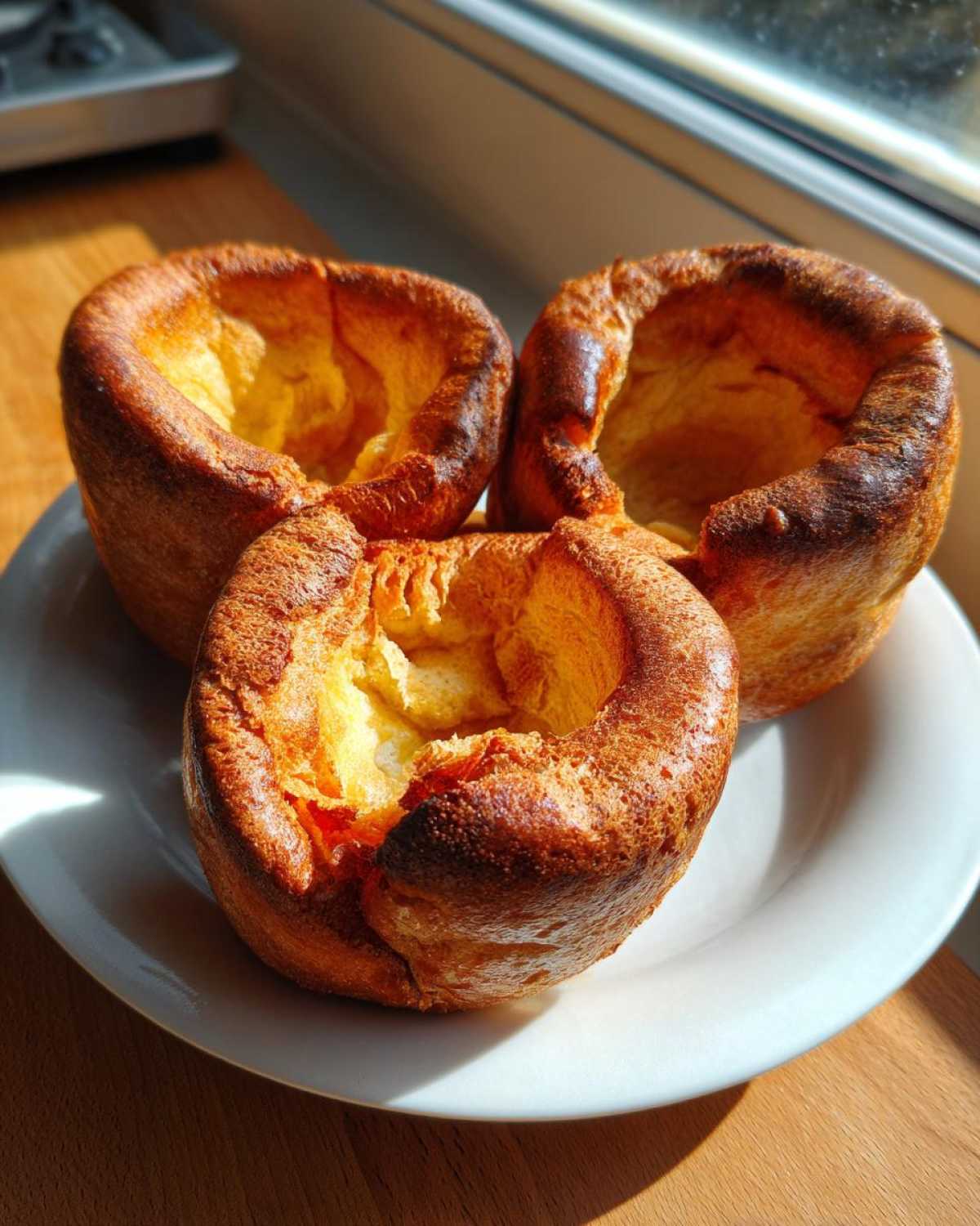 Three golden brown, tall, and fluffy yorkshire pudding pastries sitting on a white plate.