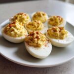 Six perfectly prepared deviled eggs arranged on a white plate, topped with creamy filling and sprinkled with paprika.