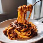 A fork lifts a generous portion of pasta coated in rich bolognese sauce from a white plate.