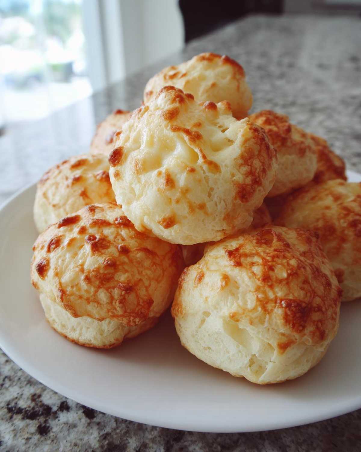 A pile of golden-brown, fluffy brazilian cheese bread rolls with melted cheese topping served on a white plate.
