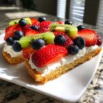 Two slices of fruit pizza sugar cookie topped with whipped cream, fresh strawberries, blueberries, and kiwi.