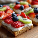 Close-up of a square fruit pizza sugar cookie topped with cream cheese frosting, strawberries, kiwi, and blueberries.