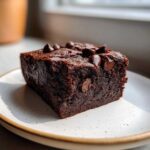 A close-up, fudgy square of black bean brownies topped with melted chocolate chips on a speckled white plate.