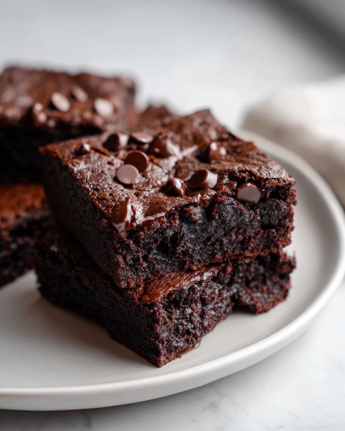 Close-up of two stacked, fudgy black bean brownies topped with chocolate chips on a light plate.