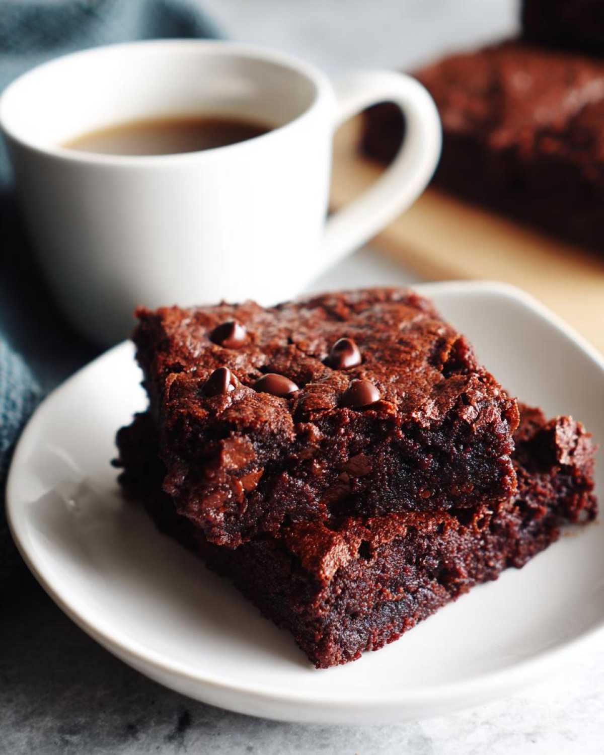 Two fudgy date brownies stacked on a white plate next to a mug of coffee.