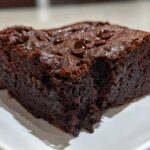 Close-up of a rich, fudgy slice of date brownies topped with chocolate chips on a white plate.