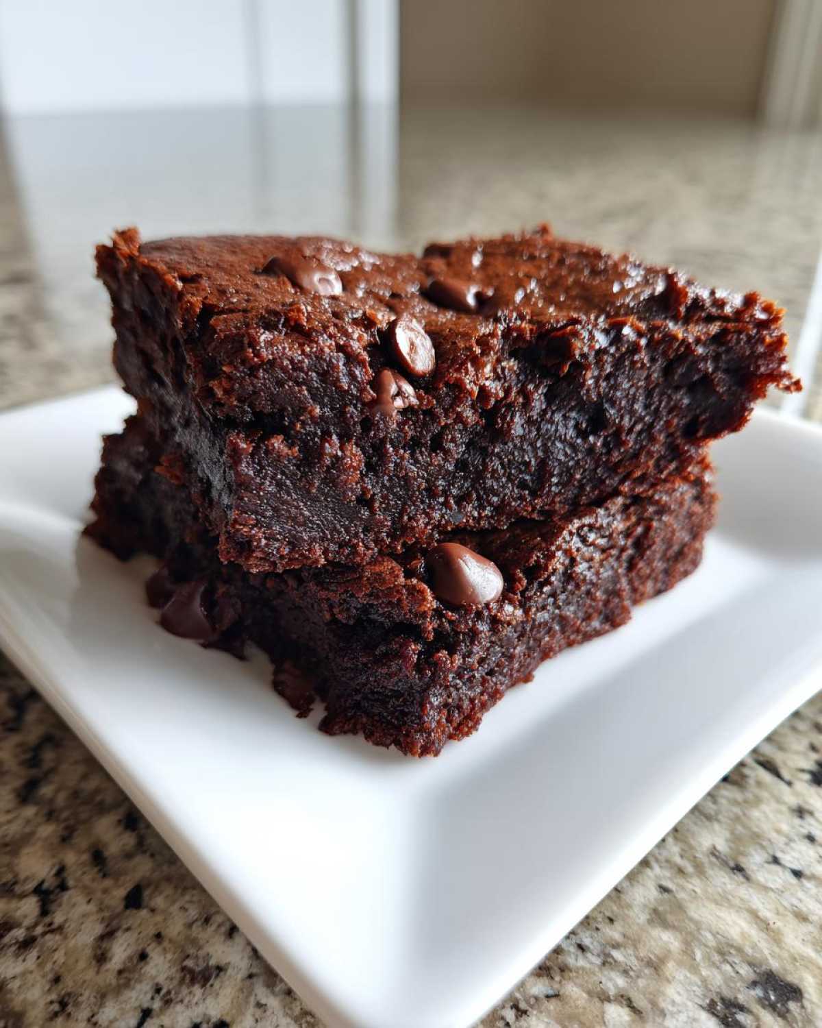 Two stacked, fudgy date brownies topped with melted chocolate chips, served on a white square plate.