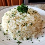 A mound of fluffy white garlic butter rice garnished with chopped parsley on a white plate.