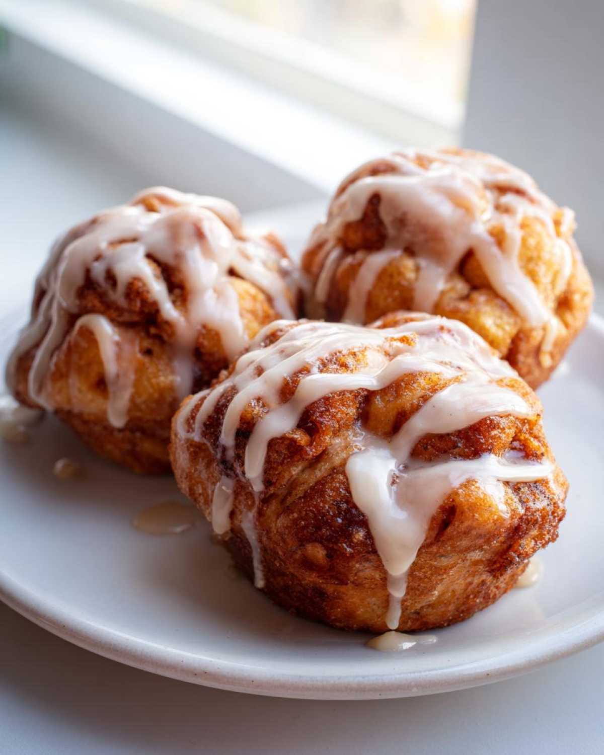 Three golden brown, round apple fritter treats drizzled heavily with white vanilla glaze, sitting on a white plate.