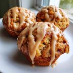 Close-up of three freshly baked apple fritter muffins topped with a thick, spiced glaze, sitting on a white plate.