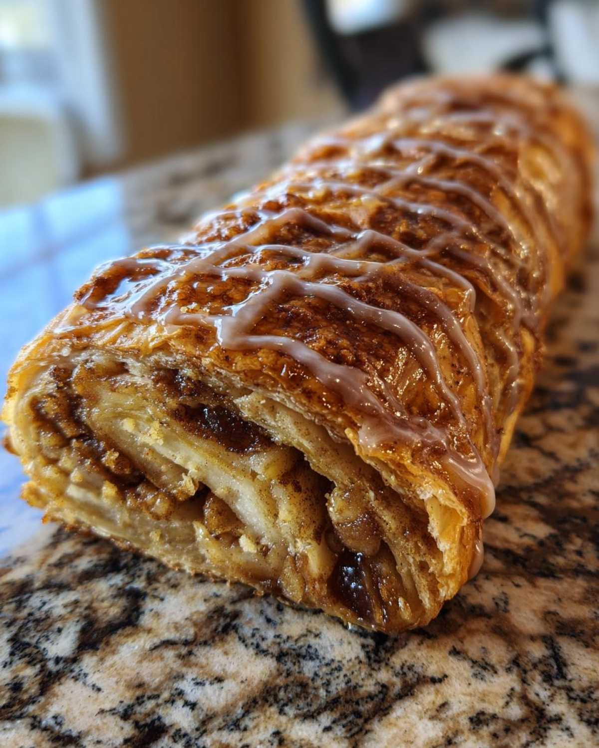 A close-up of a freshly baked apple strudel roll, drizzled with white icing, showing flaky layers and apple filling.