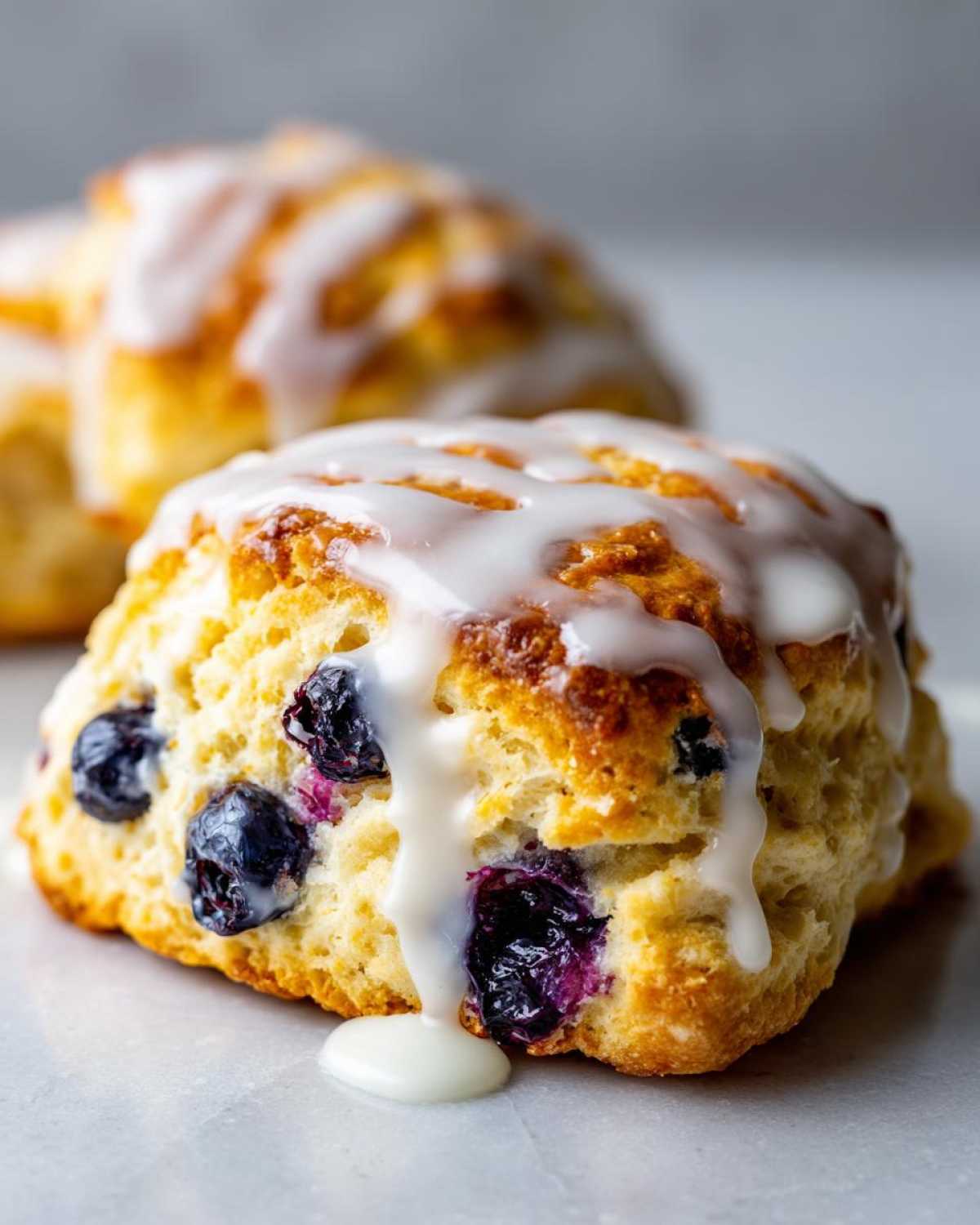 Close-up of a freshly baked, tall blueberry scone drizzled with thick white glaze, showing juicy blueberries inside.