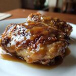 Close-up of two succulent, glazed braised chicken thighs resting in their sauce on a white plate.