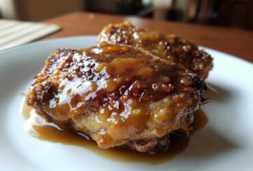 Close-up of two succulent, glazed braised chicken thighs resting in their sauce on a white plate.