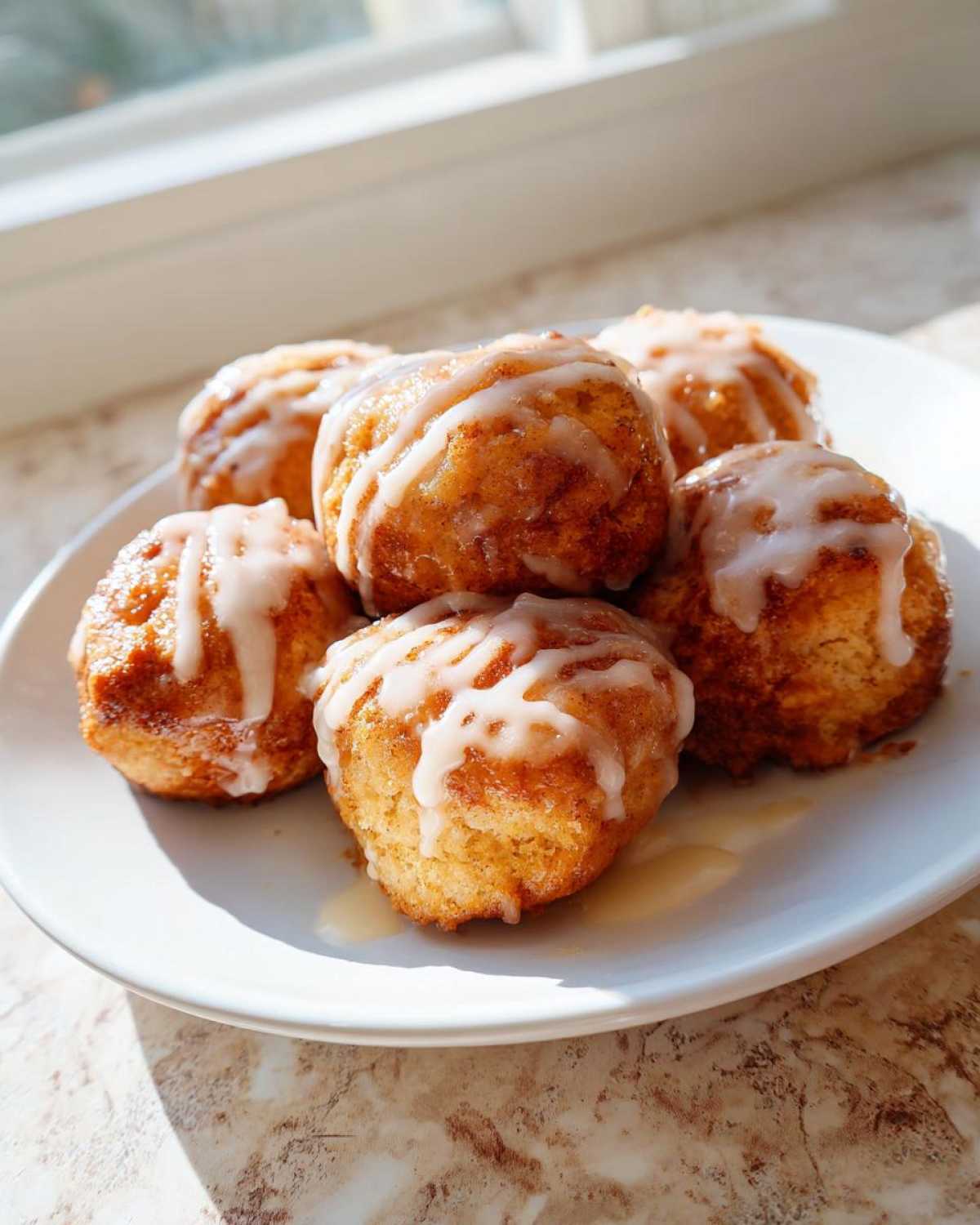 A plate of freshly baked cinnamon roll bites drizzled with white icing, sitting near a bright window.