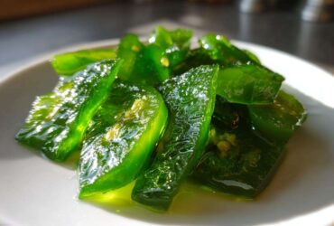 Close-up of glossy, bright green slices of candied jalapenos sitting in a light syrup on a white plate.