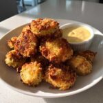 A pile of crispy, golden brown Fried Pickles served on a white plate next to a small ramekin of yellow dipping sauce.