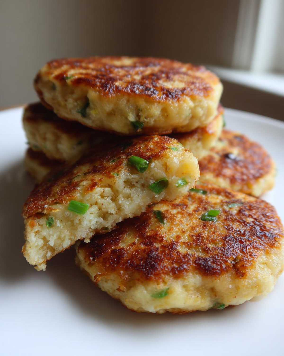 A stack of golden brown, pan-fried mashed potato cakes, one cut open to show the fluffy interior with green onions.
