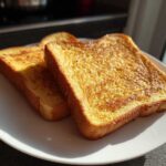 Two golden-brown slices of protein french toast stacked slightly on a white plate, catching bright sunlight.