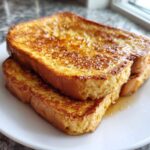 Close-up of two thick slices of golden brown protein french toast stacked on a white plate, glistening with syrup.