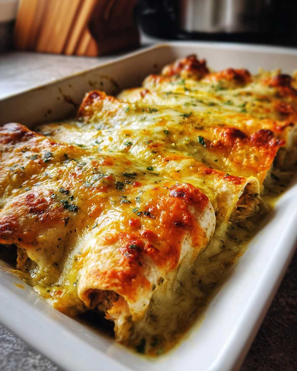 Close-up of baked green chile chicken enchiladas covered in melted, browned cheese and herbs in a white baking dish.
