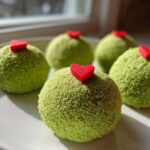 Close-up of four green, textured Grinch Oreo balls topped with small red candy hearts on a white plate.