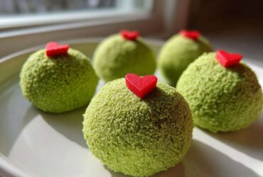Close-up of four green, textured Grinch Oreo balls topped with small red candy hearts on a white plate.