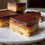 Four square pieces of homemade twix bars showing a shortbread base, caramel layer, and chocolate topping on a white plate.