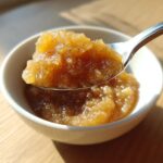 A spoonful of chunky, golden homemade applesauce being lifted from a small white bowl in bright sunlight.