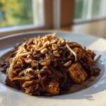 A close-up, sunlit shot of delicious homemade Pad Thai noodles topped with crushed peanuts and tofu cubes.