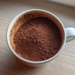 Overhead close-up of rich brown hot cocoa mix powder filling a white mug, ready for mixing.