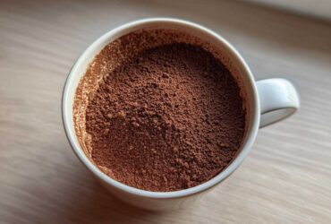 Overhead close-up of rich brown hot cocoa mix powder filling a white mug, ready for mixing.