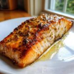 Close-up of a perfectly cooked, seasoned fillet from a baked salmon recipe resting in its juices on a white plate.