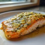 Close-up of a perfectly cooked baked salmon recipe fillet topped with garlic and herbs on a white plate.