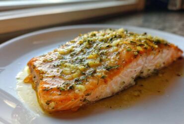 Close-up of a perfectly cooked baked salmon recipe fillet topped with garlic and herbs on a white plate.