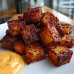 A close-up of crispy, seasoned Mexican Potatoes piled on a white plate next to a dollop of orange dipping sauce.