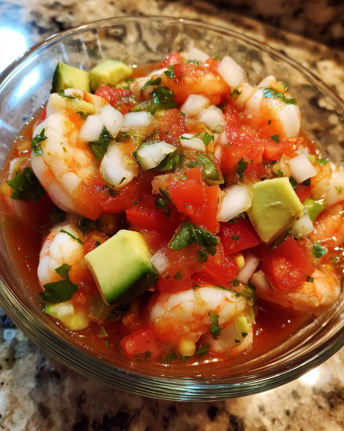Close-up view of a vibrant mexican shrimp cocktail featuring large pink shrimp, diced tomatoes, onions, avocado chunks, and cilantro in a tomato-based sauce.