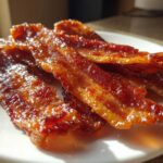 Close-up of several shiny, caramelized slices of Millionaire bacon stacked on a white plate.