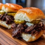 Close-up of juicy Mississippi pot roast sliders topped with melted cheese and pickles on a wooden cutting board.