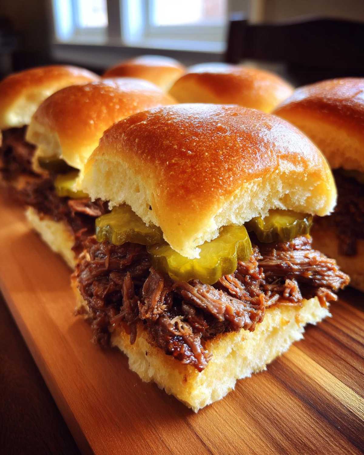 Close-up of several Mississippi pot roast sliders featuring shredded beef and pickles on soft buns, resting on a wooden board.