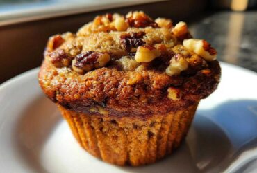 Close-up of a single, moist banana nut muffin topped generously with chopped walnuts, sitting on a white plate.