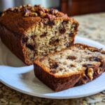 A moist loaf of date nut bread, partially sliced, topped with pecans, sitting on a white plate.