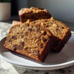 Two thick, moist slices of date nut bread featuring visible raisins and walnuts, served on a white plate.