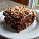 A close-up of a moist slice of german chocolate poke cake topped with gooey caramel and toasted coconut-pecan frosting.