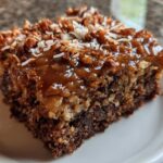 Close-up of a moist slice of german chocolate poke cake topped with gooey caramel, pecans, and coconut.