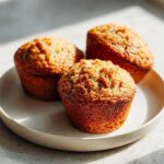 Three golden brown honey bran muffins with crumbly tops resting on a light speckled plate.