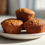 Three perfectly baked, moist honey bran muffins stacked slightly on a small white plate in bright sunlight.