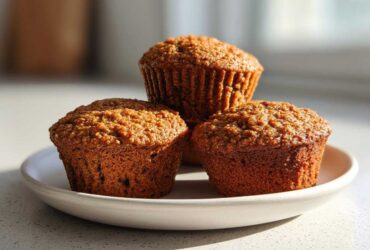 Three perfectly baked, moist honey bran muffins stacked slightly on a small white plate in bright sunlight.